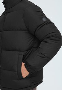 Svart puffjakke med polstret design, elastiske mansjetter, to sidelommer og glatt tekstur. Har en rund svart logo på ermet.
