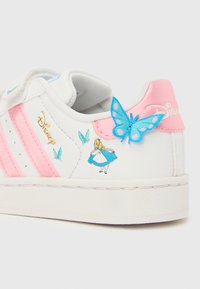 Pantof sport alb cu dungi roz, logo Disney, grafică Alice în Țara Minunilor și un fluture albastru pe partea din spate a călcâiului, pe fundal alb.