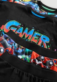 Schwarze T-Shirts mit "GAMER"-Grafik in mehrfarbigen Farbspritzern, kombiniert mit bunten gemusterten Shorts mit Gamecontroller-Designs.