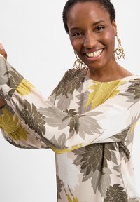 Lächelnde Frau mit blumengemustertem Blusenoberteil mit langen Ärmeln und goldenen, geometrischen Blattschmuckohrringen vor einfacher Hintergrund.