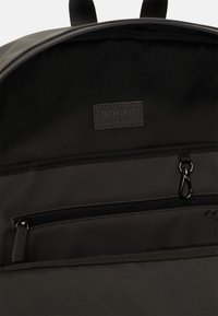 Interno di uno zaino nero con una superficie liscia opaca, patch del logo e una tasca con cerniera dotata di tiralampo in metallo.