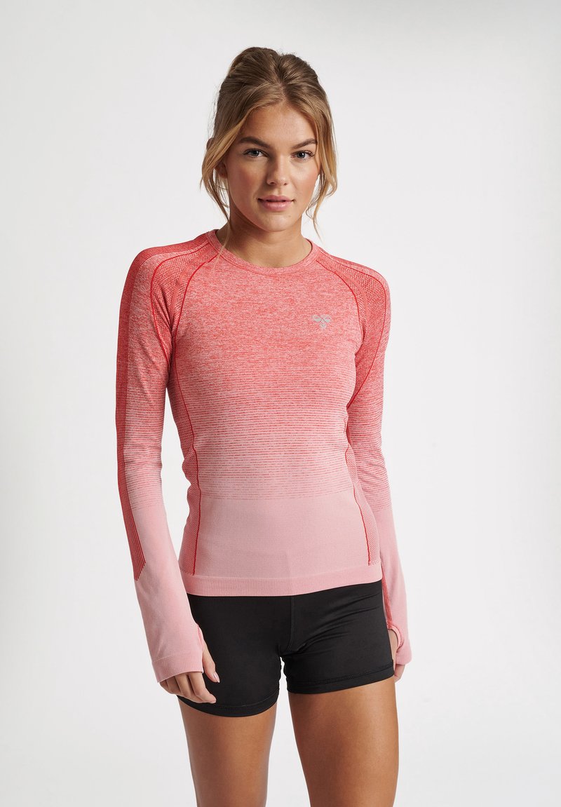 Junge Frau trägt ein eng anliegendes langärmliges Sporttop in Pink mit Farbverlauf und schwarze Shorts, steht vor einem schlichten hellen Hintergrund.