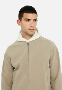 Beige kordajakke med lynlås foran, der har en hvid hætte. Stoffet har en ribbet tekstur og en afslappet pasform.