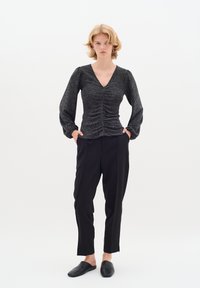 Top nero a maniche lunghe con scollatura a V e dettaglio arricciato in vita, abbinato a pantaloni neri a sigaretta e scarpe slip-on. Texture liscia.