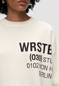Krämfärgad sweatshirt i mjukt tyg, med djärv svart grafisk text och siffror på framsidan. Förlängda ärmar och ribbad halsringning.