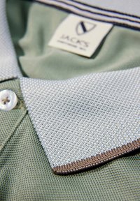 Grön polo-shirt med en strukturerad tyg, med en ljusgrå krage med brun kant och en vit knapp. Etikett synlig inuti kragen.