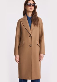 Kobieta w długim płaszczu w kolorze camel, nosząca niebieską koszulę z guzikami, w okulary przeciwsłoneczne w musze oraz złote kolczyki w kształcie kół.