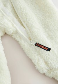 Baltas pūkuotas zip-up drabužis su minkštu tekstūru; turi centrinius užtrauktukus ir juodą etiketę, pažymėtą "SUPERDRY" šone.