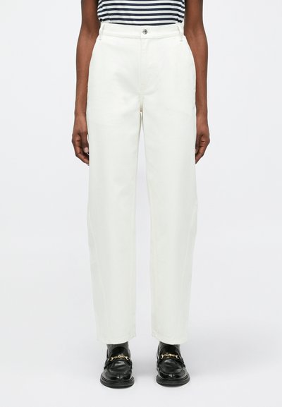 Personne portant un pantalon droit blanc taille haute, des mocassins noirs avec des chaînes dorées, des chaussettes grises et un haut à rayures noir et blanc, debout.
