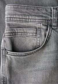 Close-up van een grijze denim jeanszak met stikseldetails en een metalen rivet op de stoftextuur.