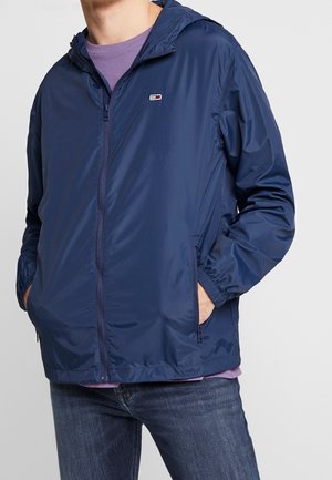 Mand iført en marineblå hættejakke med lynlås og hænderne i sidelommerne, over en lilla skjorte og blå jeans.