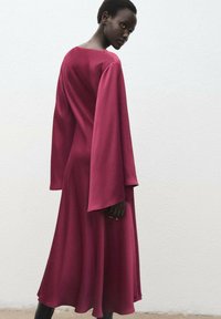 Robe fluide à manches longues de couleur bourgogne profond, fabriquée en tissu lisse avec un décolleté large et une silhouette évasée, atteignant le milieu du mollet.