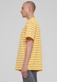 Junger Mensch mit kurzen Haaren, der ein gelb-weiß gestreiftes T-Shirt und graue Hosen trägt, im Profil vor einem schlichten Hintergrund stehend.