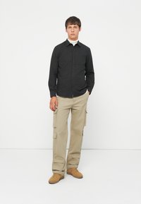 Camicia nera con bottondown, maniche lunghe, in tessuto misto cotone; abbinata a pantaloni cargo beige con tasche; sneakers color nocciola completano l'outfit.
