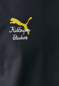 Svart stoff med en gul puma-logo og hvit "KidSuper Studios"-tekst. Glatt tekstur, enkel design uten ytterligere mønstre.