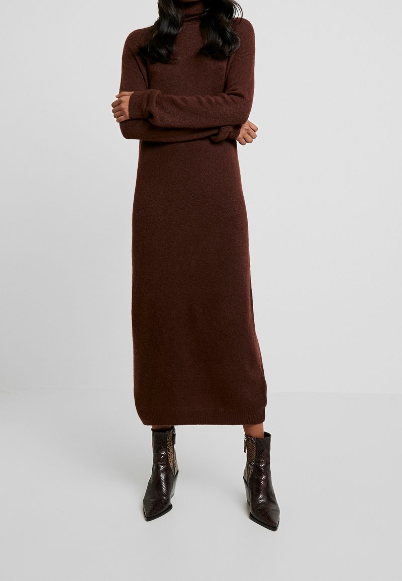 Femme portant une longue robe-pull marron à col roulé avec des manches longues et des bottines marron foncé à bout pointu.