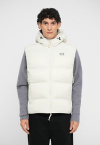 Puffy cream-colored vest with a high collar and hood. Features zippered side pockets and a small logo patch on the chest.