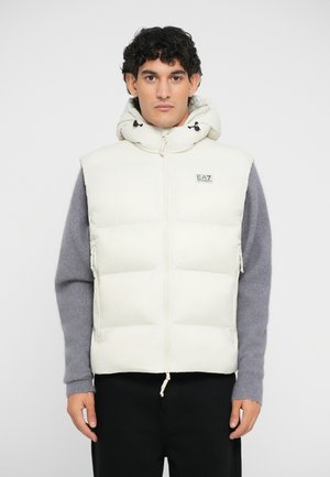 Puffy cream-colored vest with a high collar and hood. Features zippered side pockets and a small logo patch on the chest.