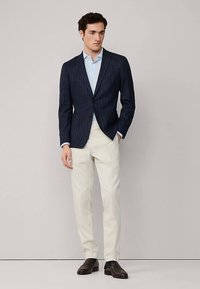 Mornarska plava sako jakna na prugice uparena sa svijetloplavom košuljom, krem hlačama i tamnim cipelama. Sako ima kroj po mjeri i jedan gumb.