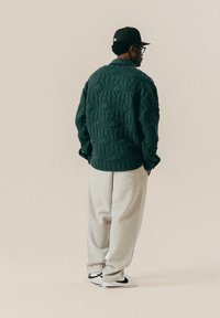 Άνδρας φορώντας ένα σκούρο πράσινο καπιτονέ μπουφάν, ανοιχτού γκρι φόρμες, λευκά αθλητικά παπούτσια, μαύρο καπέλο και γυαλιά, στέκεται με τα χέρια στις τσέπες κοιτώντας προς τα πίσω.