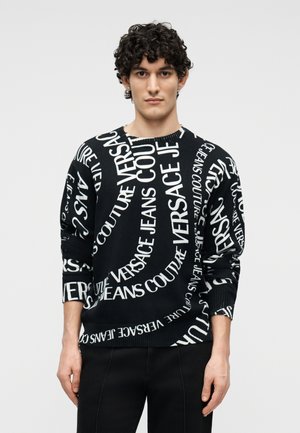 Tânăr cu păr creț și închis la culoare, purtând un pulover negru acoperit cu text mare alb Versace Jeans Couture, stând în fața unui fundal simplu și deschis la culoare.