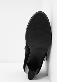 Semelle de chaussure à talon noire avec motif de bande de roulement texturé, en angle contre une surface blanche à coins.