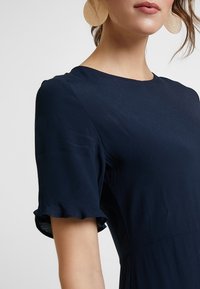 Žena v tmavě modrých šatech s krátkými volánkovými rukávy a velkými kulatými zlatými náušnicemi na jednobarevném pozadí.