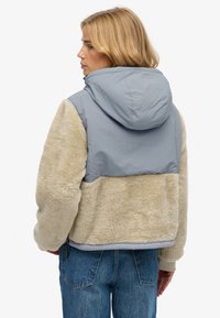 Kunstpelzjacke mit beigem, strukturiertem Stoff und einem hellgrauen Nylonpanel sowie Kapuze. Verfügt über einen Reißverschluss und gerippte Bündchen.