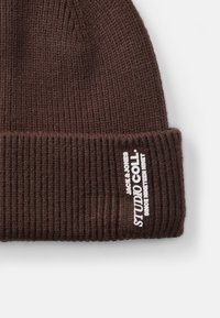 Bruine gebreide beanie met ribbelstructuur, voorzien van een omgeslagen boord en witte brandingtekst aan de voorkant.