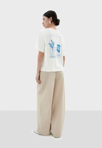 Witte T-shirt met blauw grafisch ontwerp met botanische elementen. Gecombineerd met beige wijdere broek, van een soepele stof en een relaxte pasvorm.