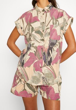 Vrouw draagt een kortarmig hemd met knopen en bijpassende korte broek met groot paars-groen bloemenpatroon op een lichte achtergrond.