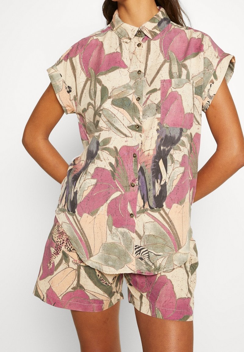Femme portant une chemise à manches courtes boutonnée et un short assorti avec un grand motif floral violet et vert sur fond clair.