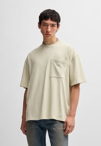 Světlé béžové oversized tričko vyrobené z bavlny, s kapsou na hrudi a minimalistickými švy. Nosené s vybledlými modrými džíny.