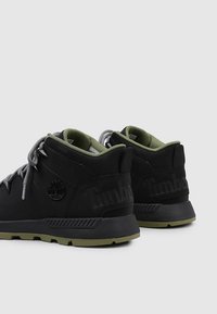 Zapatillas negras de corte medio con cordones grises y acentos verdes. Presentan un detalle de logo en el talón y un material negro texturizado. Suela de goma.