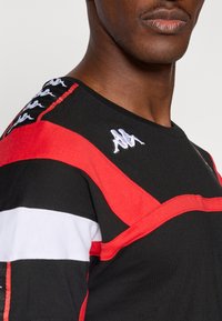 Μαύρο και κόκκινο κοντομάνικο πουκάμισο με λευκές ρίγες, διαθέτοντας ένα υ�φασμένο λογότυπο και κοντράστ ραφές. Λείο ύφασμα με αθλητικό σχεδιασμό.