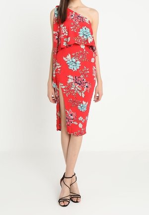 Kvinde iført en rød one-shoulder midi kjole med blå og pink blomsterprint og en højtudskæring på låret, kombineret med sorte sandaler med stropper.