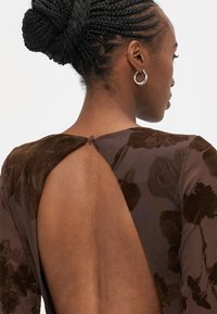 Καφέ φόρεμα με λουλουδάτο σχέδιο και βαθιά κομμένη πλάτη. Διαθέτει λεία υφή και κλείσιμο με κουμπιά στον λαιμό. Ορατά ασημένια κρίκοι σκουλαρίκια.