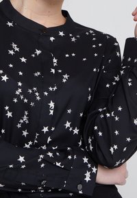 Svart skjorta med ett stjärnmönster i vitt, med knappstängning fram, krage och långa, något rynkade ärmar. Mjuk, slät tyg.