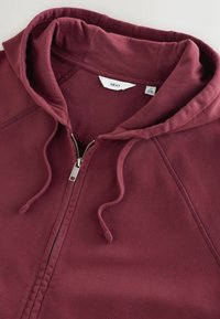 Sweat à capuche bordeaux à zip avec cordons et capuche, présenté à plat avec l'étiquette de la marque "NEXT" visible et une étiquette de taille moyenne.