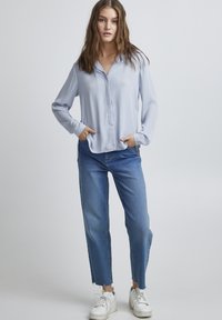 Blusa azzurro chiaro a maniche lunghe con colletto, abbinata a jeans in denim a gamba dritta e cropped con fessure laterali e sneakers bianche.