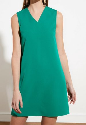 Frau trägt ärmelloses, knielanges grünes Kleid mit V-Ausschnitt und goldenen Creolen, steht vor weißer Wand.