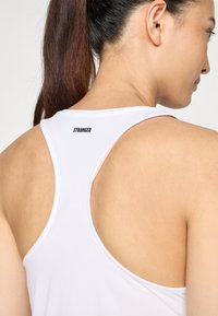 Valge spordiline tank top, millel on selja taha ristatud disain. Ülal taga on must "STRONGER" logo. Sile ja kerge kangas.