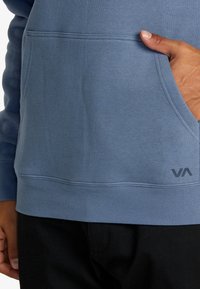 Felpa con cappuccio in tessuto blu, dotata di tasca frontale e bordo a coste. Presenta un logo nero posizionato vicino in basso a destra.