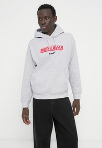 Γκρι βαμβακερή φούτερ με κουκούλα και μπροστινή τσέπη, με κόκκινο γραφικό κείμενο "METAL GEAR" και λογότυπο. Συνδυασμένη με μαύρα τζιν.