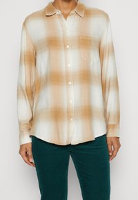 Camisa de manga larga a cuadros en tonos beige y blanco, con botón frontal, cuello y bolsillo en el pecho, hecha de una tela texturizada.