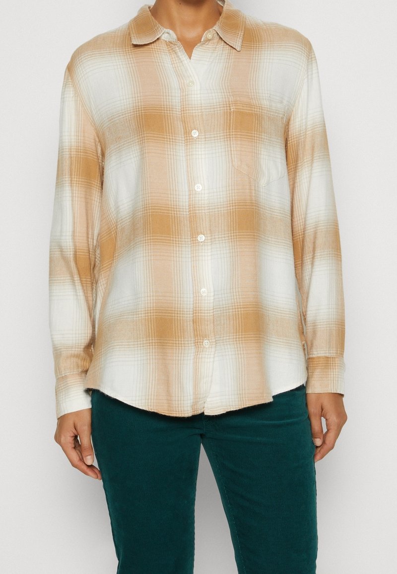 Camisa de manga larga a cuadros en tonos beige y blanco, con botón frontal, cuello y bolsillo en el pecho, hecha de una tela texturizada.