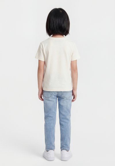 Otrok s kratkimi črnimi lasmi stoji obrnjen stran, oblečen v preprosto belo majico, svetlo modre kavbojke in bele superge na belem ozadju.