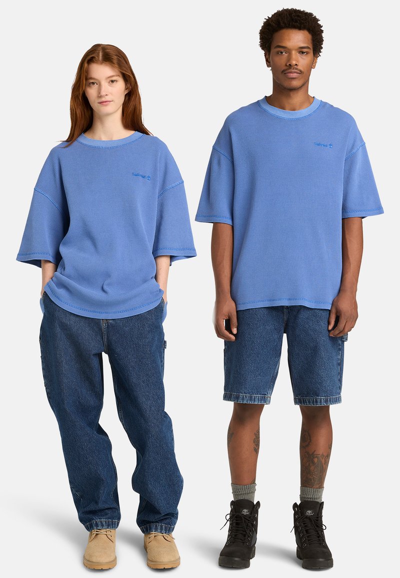 Deux mannequins portant des hauts bleu oversize à manches courtes ; l'un avec un jean bleu ample et des chaussures marron clair, l'autre avec un short en denim, des chaussettes grises et des bottes noires.