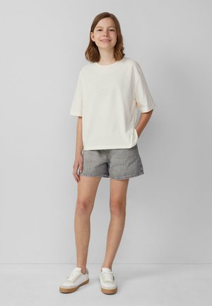 Lachend staande meisje in een wit oversized T-shirt, grijze denim shorts en witte sneakers tegen een effen lichtgrijze achtergrond.