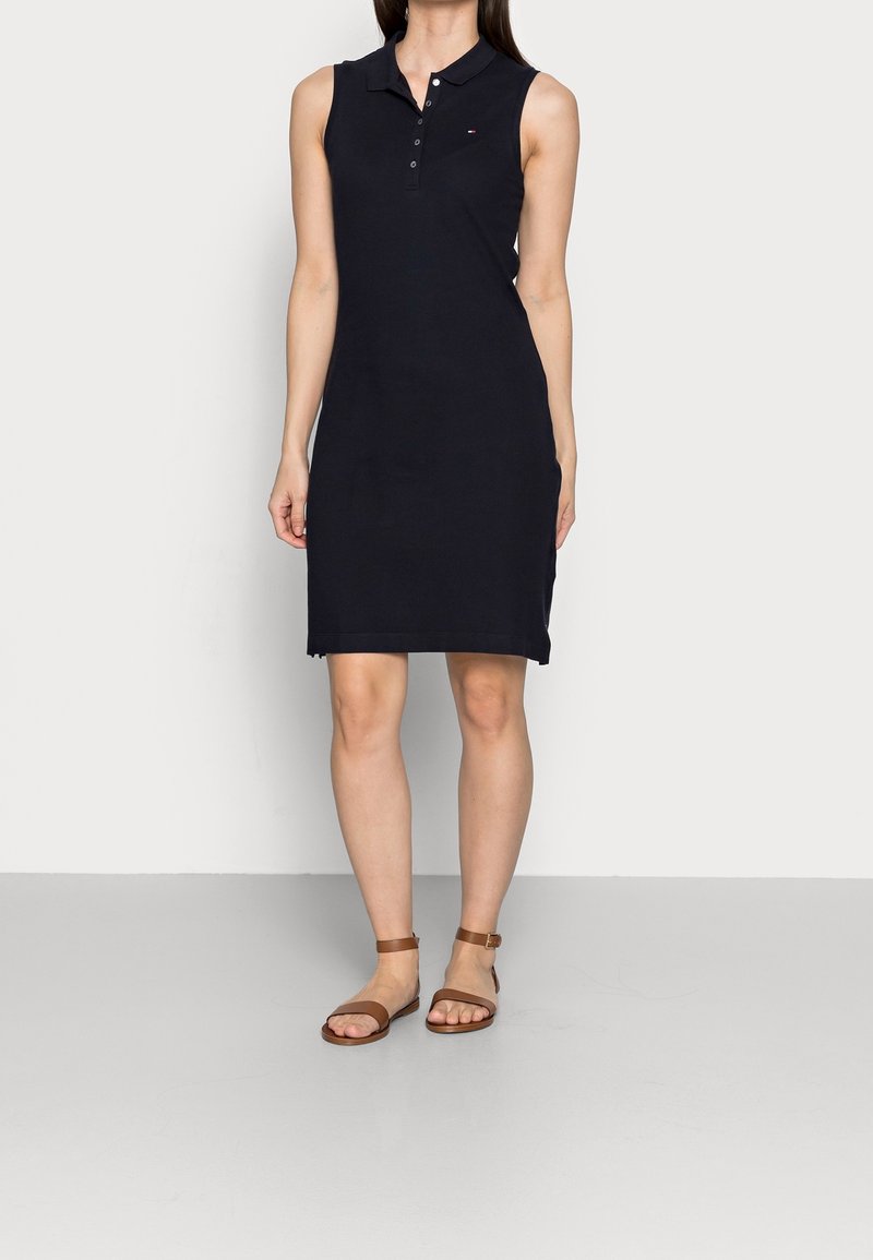Femme portant une robe polo noire sans manches et des sandales plates marron à bride à la cheville, debout devant un fond clair uni.